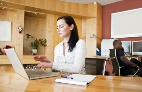 Businesswoman working on laptop with male partner sitting in background at office